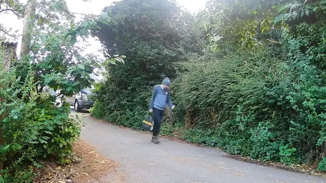 Roy Barclay is walking down a country lane wearing a blue beanie hat, a blue jumper, dark trousers and walking boots. He is carrying a black plastic carrier bag. On either side of the lane are green shrubs and trees.