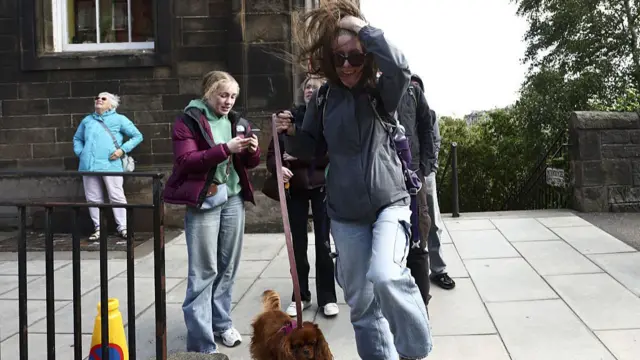 A woman in a grey jacket and light-blue jeans moved her hair away from her face after a strong gust of wind. In her right hand, she's holding a pink lead connected to a small brown dog. A young blonde girl stands behind her, to the right, in a purple jacket, green hoodie and jeans, another woman and a man to her left