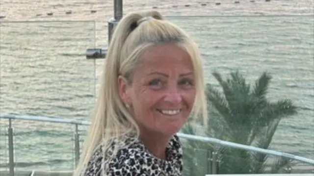 Anita Rose is wearing a leopard print top and has her long blonde hair in a high ponytail. She is looking at the camera and smiling her. She appears to be sat on a rooftop and in the background is a calm sea and palm trees