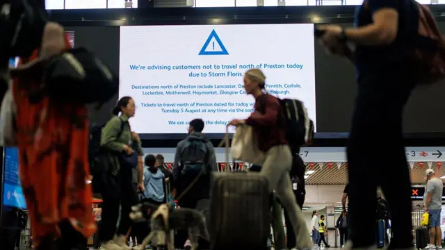 A travel warning on a screen at a station, advising people not to travel north of Preston, as people walk in the foreground with luggage