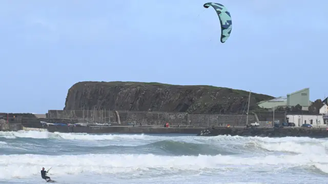 A man is seen kite surfing on white cresting waves in front a rock face