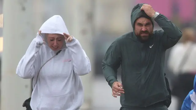 A man and woman walking in the rain, hands holding hoods as rain pours.
