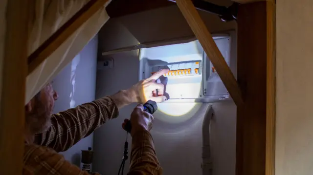 A man in a long-sleeved shirt holds up a torch to an electricity meter during a power outage