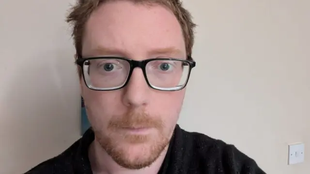 A man with red hair and glasses in a black t-shirt against a cream wall