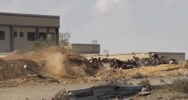 Screengrab from a verified video, filmed near an aid distribution site near Nuseirat in Gaza, which shows dirt in the air beside a road following gunfire. Beyond the road are people hiding behind a large mound, with buildings behind them.
