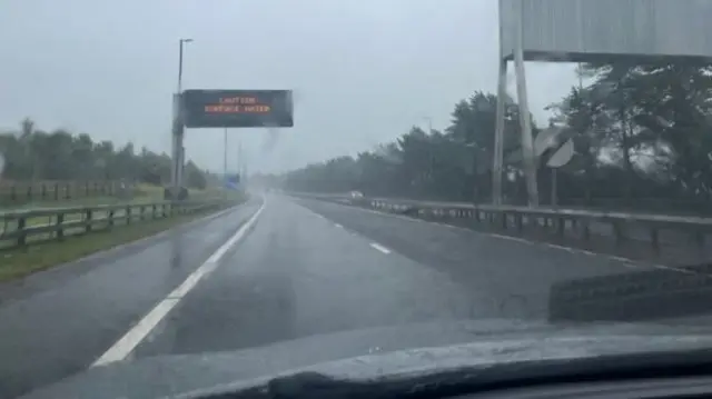 Motorway with sign saying 'Caution, surface water' - rain pouring onto road and car windshield.