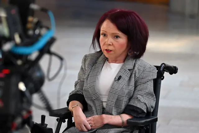 Pam Duncan-Glancy has dark shoulder-length hair and his wearing a black and white checked jacket over a white top. She is sitting in a wheelchair