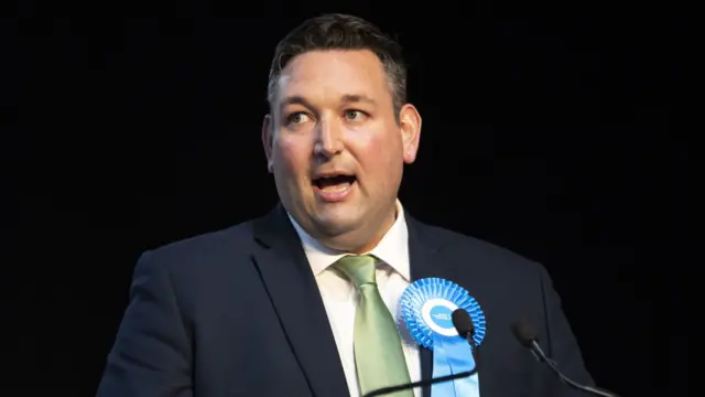 Miles Briggs standing against a black background, wearing a dark suit over a white shirt and pale green tie and a blue rosette on his lapel.