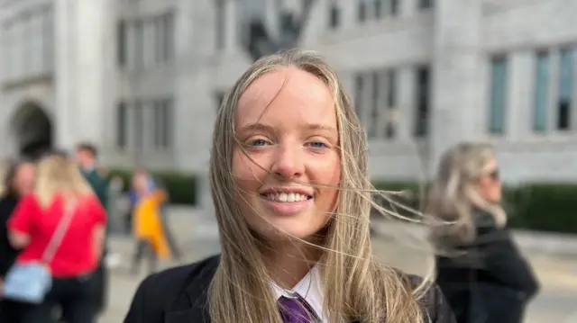 Katy McDonald smiling at the camera. She has blonde hair partially covering her face and is wearing a black blazer over a white shirt with a purple car.