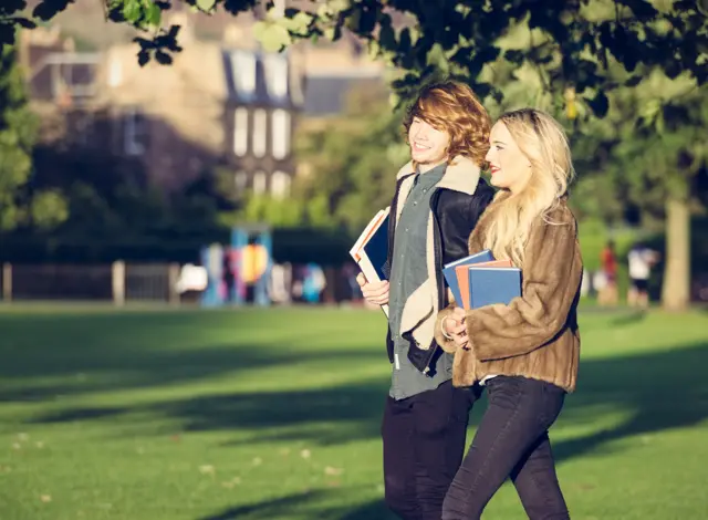 Two teenage students holding books as they walk through a city park