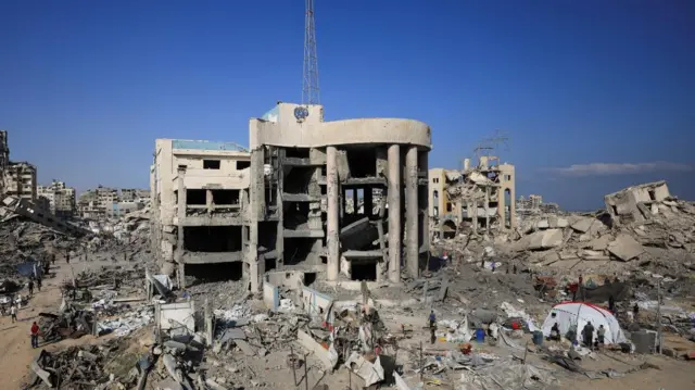 The shell of a bombed-out building, with a distinctive curved facade, stands surrounded by rubble