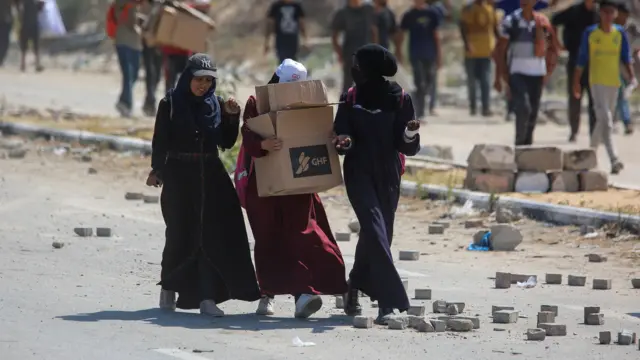 Women holding a GHF box walk down a road in Gaza