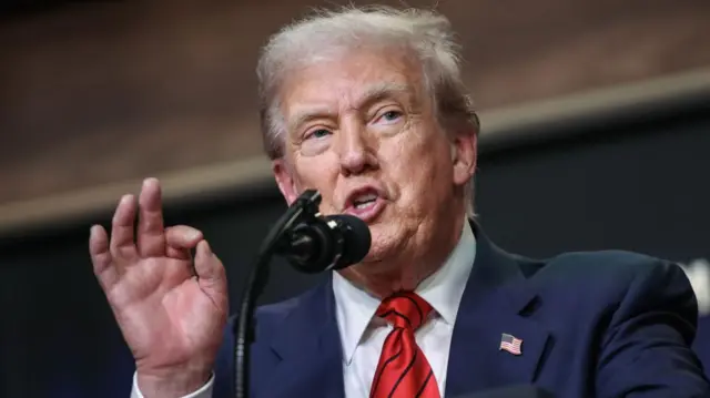 A close up of Trump speaking at a lectern. He pinches his forefinger and thumb together in gesture as he speaks