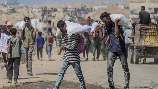 Two men carry sacks on their backs as they walk through sand, in the background are more people walking with some carrying bags on their shoulders.
