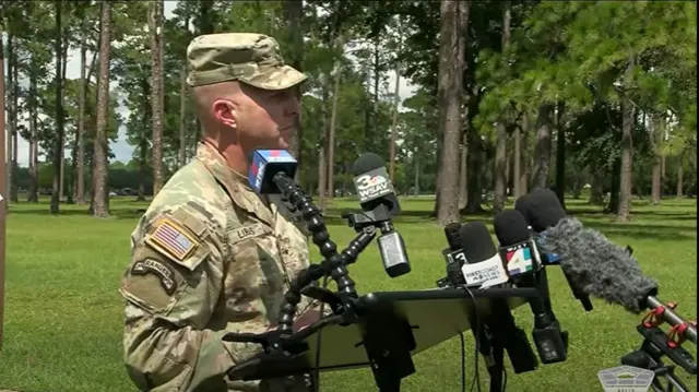 A man in Army uniform speaks to press from behind some microphones.
