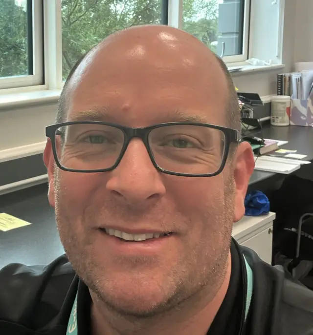 A balding man smiles as he looks at the camera inside an office.
