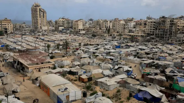 Wide shot of a large encampment of shelter tents in Gaza City, damaged buildings in the background