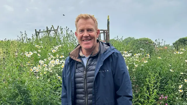 Adam Henson, wearing all navy blue - blue jeans and a blue waterproof - stands in front of a tall bed of wildflowers