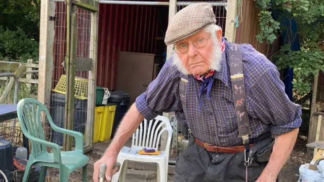 A man with white hair and white sideburns. He is wearing a flat cap and a checked shirt with braces which have horses on them. He is holding a crutch and wears glasses.