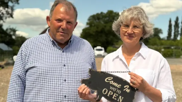 Peter and Alison Themans from Wenlock Edge Farm Shop are standing on the empty ground where the building previously stood before the fire. Alison is holding a metal pig-shaped sign saying 'come in, we're open'. Peter has receding grey hair and is wearing a checked blue and white shirt. Alsion has short white/grey hair and is wearing a white shirt.