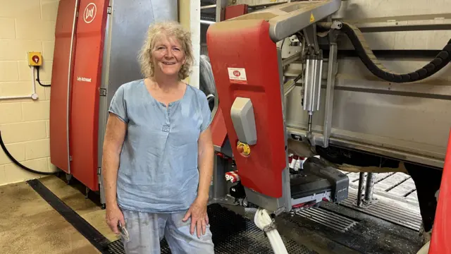 A middle aged woman with mid-length blonde curly hair, a pale blue top and light blue, dirty jeans stands in front of a red and grey robotic device on a farm