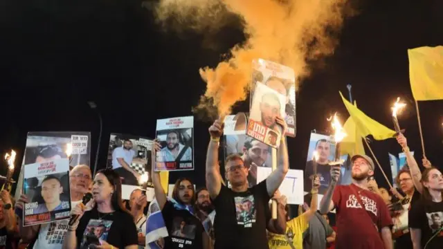 A group of people holding posters of hostages, some hold flares that are on fire and have orange smoke, others hold up yellow flags