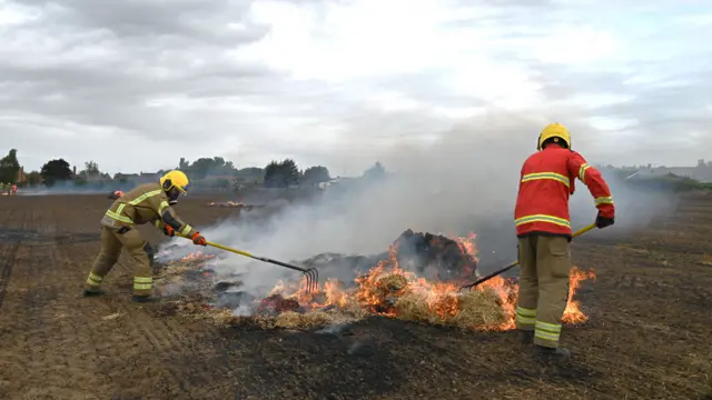 Two firefighters in head-to-toe protective gear with yellow reflective strips and yellow helmets are tackling a fire in the middle of a farm field. They are using rakes on hay that is consumed by flames.