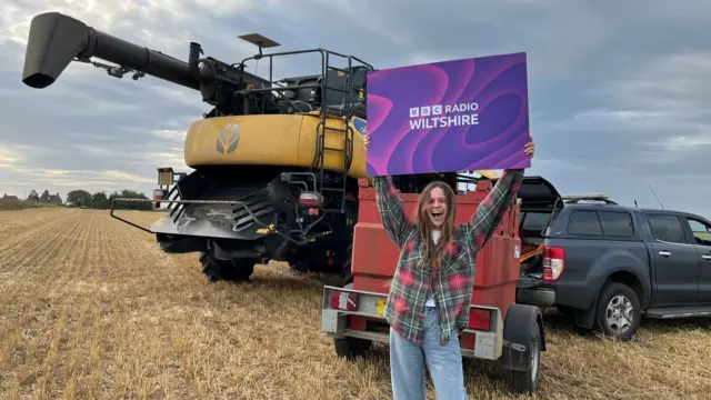 Gabrielle Aplin holds a purple BBC Radio Wiltshire sign. She is wearing jeans and a plaid shirt.