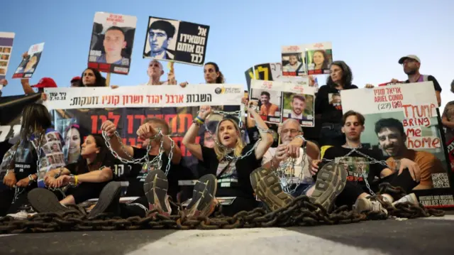 Six people (three men and three women) sitting on the ground with chains around them and their hands as a group of people holds up signs and placards behind them