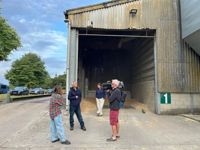 A man in dark jeans and a blue hoodie holds a microphone and looks towards a man holding a camera. Two other members people are standing out of shot, with the entrance to a grain store visible in the background
