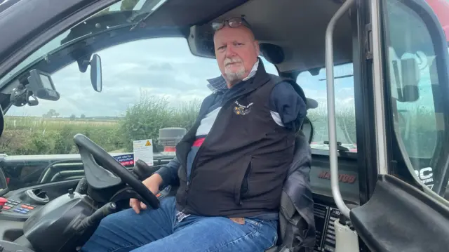 A bald man with a beard sits inside a tractor. He is wearing jeans and a gilet.
