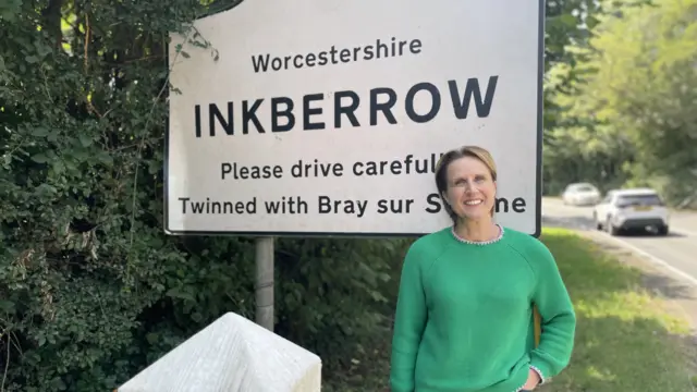 A woman with short blonde hair, wearing a green jumper and jeans, smiles in front of a large white road sign saying ‘Worcestershire INKBERROW Please drive carefully’