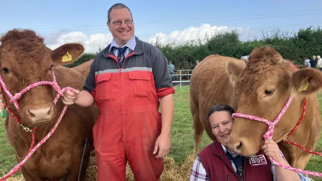 A man is stood up holding a rope attached to a large brown cow on his left. Next to him is his wife. She is crouched holding rope attached to a cow which is resting its head on her right shoulder.