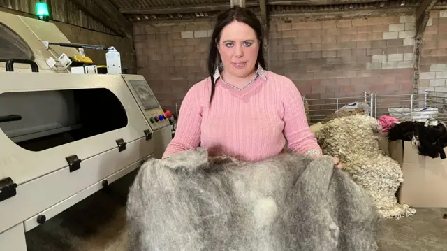 Lucinda Bird is in a brick farm building holding a grey blanket of wool up to the camera. She has long black hair and is wearing a pink jumper. She is smiling slightly.