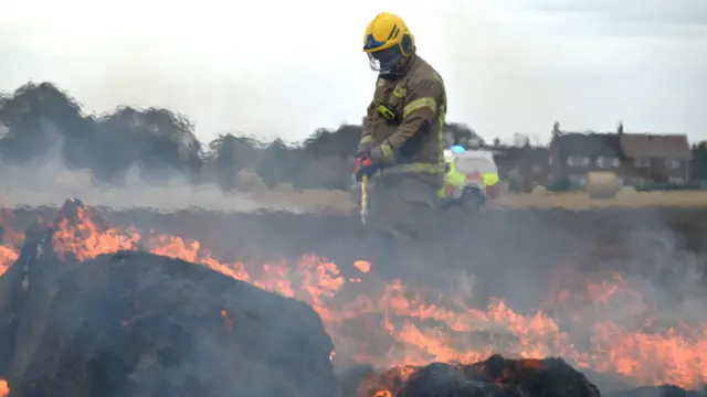 A firefighter in brown protective gear wih yellow reflective strips is raking a large fire in a farmer's field. The firefighter has a yellow helmet with a visor. Bales of hay are on fire, consumed by orange flames