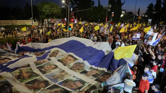 A large Israeli flag with lots of headshots of people on it, who are the hostages. Around the huge flag is a large crowd of people, who hold little yellow flags and Israeli flags.