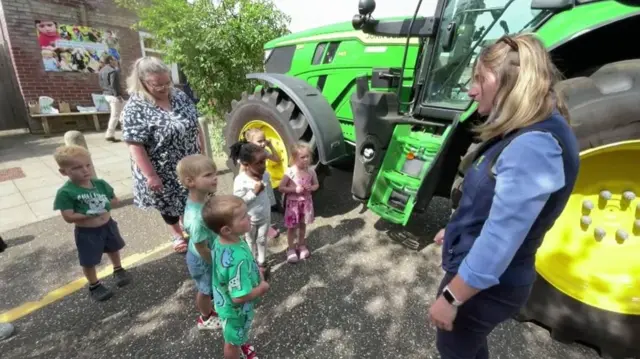 Six young children are standing with a teacher, looking at a farmer with long blonde hair. She is standing next to a green tractor with yellow in its wheels.