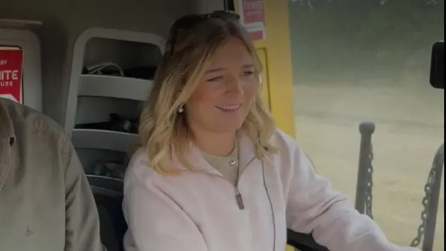 Harriet has blonde hair, pearl earrings and a pink zip jumper. She is inside a combine vehicle, next to the window, with a field visible behind her.