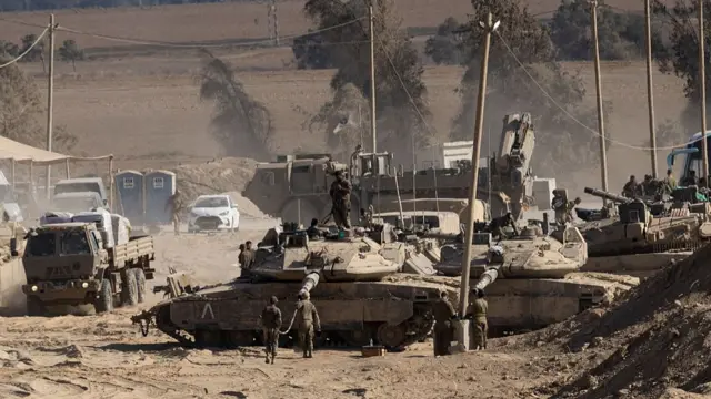 Israeli soldiers in full military gear stand next to and on top of tanks as military vehicles drive by on a dusty road near the border with the Gaza Strip