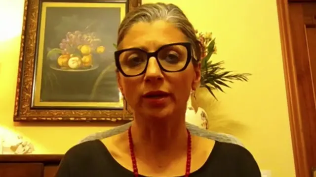 a woman wearing glasses sits in a room with a painting and lamp behind her