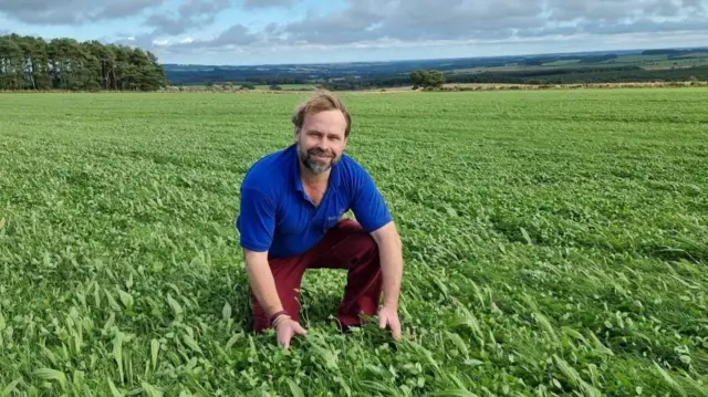 Simon Bainbridge is kneeling in a field of pasture and smiling at the camera. He has a short beard and short brown hair - and is wearing a blue shirt and red bottoms. In the background are the expansive fields of the countryside