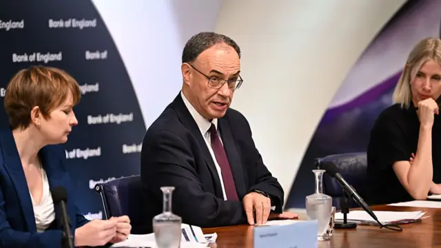 Clare Lombardelli (L) and Andrew Bailey (C) sit down at a wooden table, Bailey answering a question during a press conference