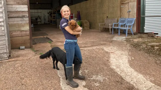 A young woman with blonde and red hair tied back is standing in a farm yard. She is wearing jeans, a t-shirt and wellington boots and is holding a brown dachshund. There is a larger black dog peeping its head through her legs. The woman is laughing.
