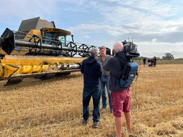 A man with a camera crew, filming two men in a field. There is a combine harvester behind them.