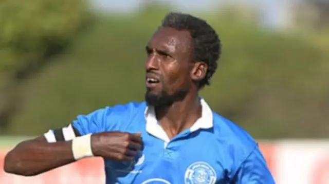 A man on a football pitch which is blurred in the background. He wears a blue football top and holds one arm in a fold to his chest. He looks to  his side