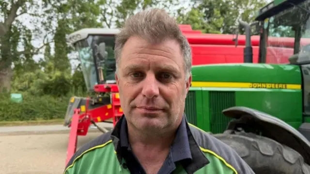 Henry has grey/brown hair and is wearing a green and black top with yellow piping. There is a large red piece of farm machinery behind him and a green John Deere tractor.