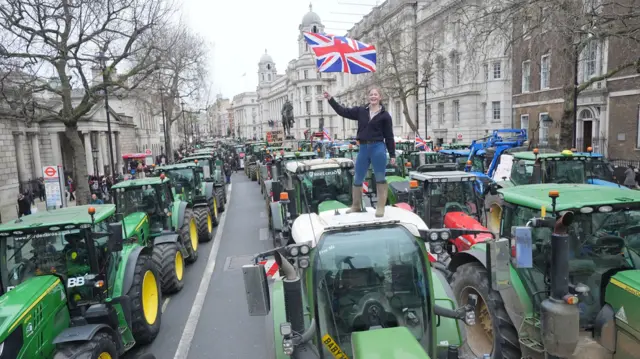Dozens of tractors parked in Whitehall as part of a protest - on top of one is a young woman wearing wellies who is waving a Union Jack