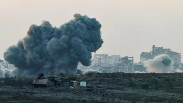 A huge plume of smoke rises from the ground following an explosion. In the background there are ruined buildings and more smoke. In the foreground is scrub land with some debris on it