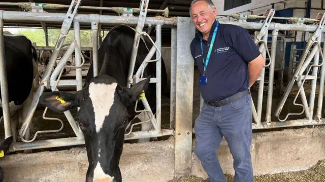 A man standing next to a black and white dairy cow. He has short grey hair and is wearing a navy blue polo shirt and blue jeans. He Is looking ahead and smiling.