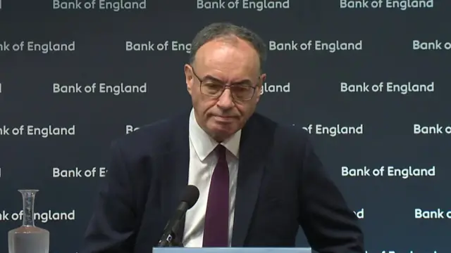 Andrew Bailey, wearing a dark suit and burgundy tie, sitting with microphone in front of him and a Bank of England branded backdrop behind him